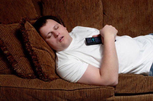 A young man sleeps on a couch, with a TV remote control firmly gripped in his hands.